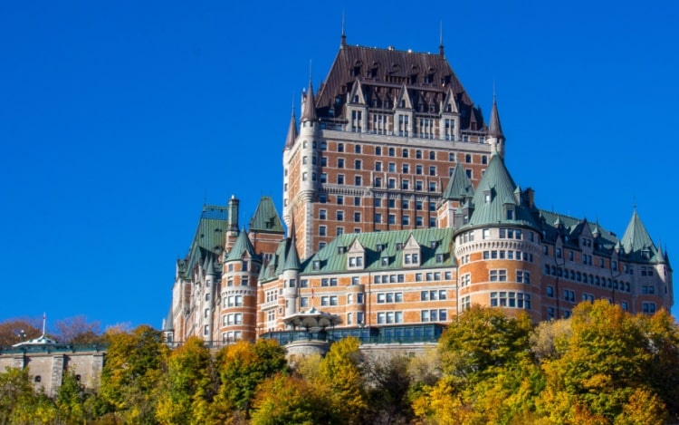 A large castle-like hotel with an orange-brown exterior and green pointed roof sits behind a row of trees under a clear blue sky.