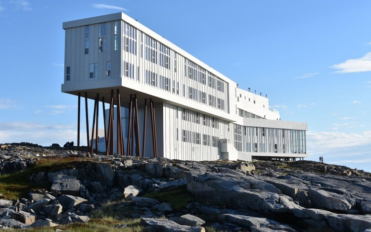 A large white building perched on brown stilts above rocks.