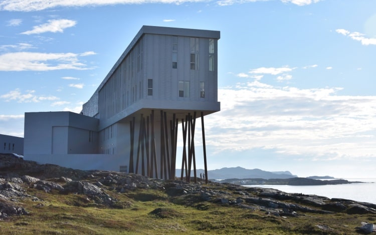 A grey building on stilts on rocky ground, with the sea visible in the distance.