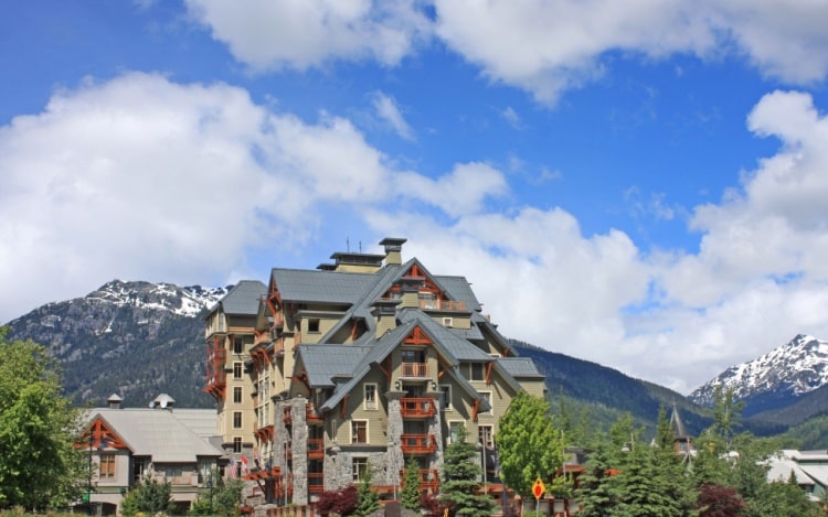 A grey and brown ski lodge building near snow-capped mountains and green trees.