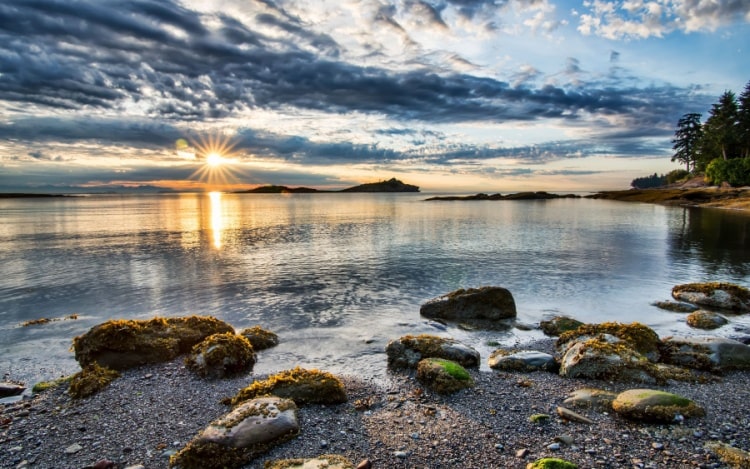 A photo of the sea taken from a rocky shore, with blue sky, dark clouds, and a bright yellow sun in the distance.