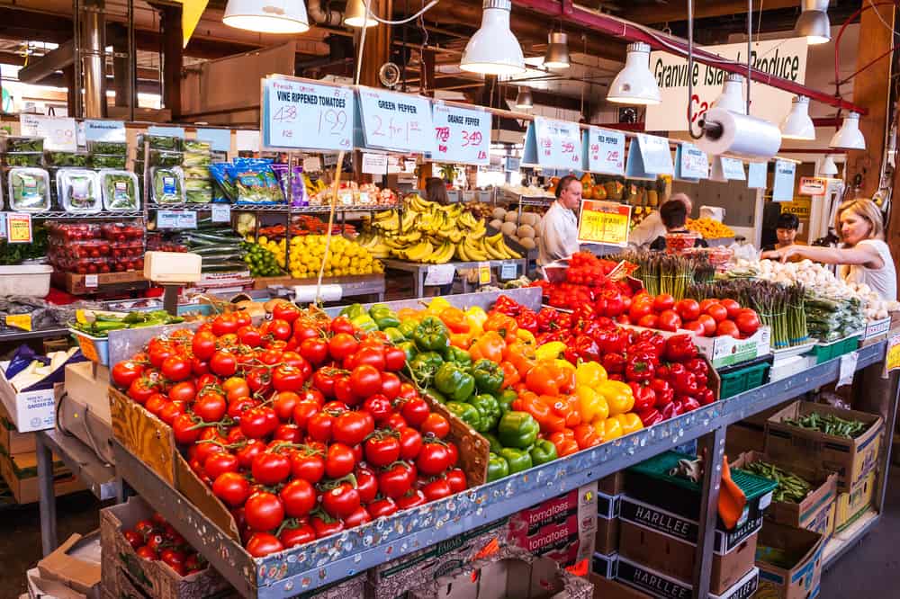 Public market with stalls offering piles of tomatoes, peppers, bananas and other fruit and vegetables to people walking by