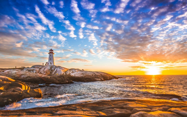 Peggy’s Cove at sunset. A lighthouse stands on rocks next to the sea, with the sun setting in the distance over the water.
