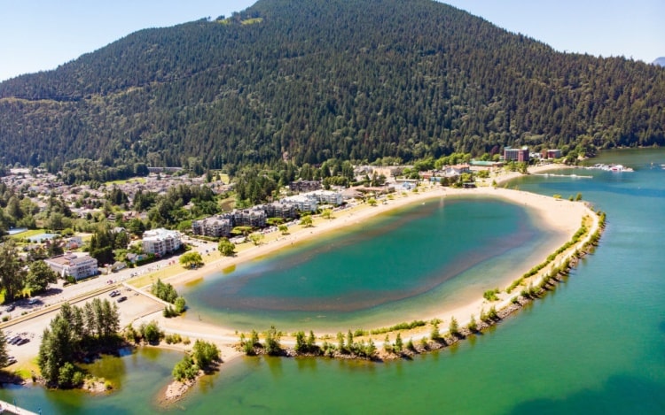 An aerial view of Harrison Hot Springs’ shoreline, with green water, sandy shoreline, buildings and a tree-covered hill in the background.