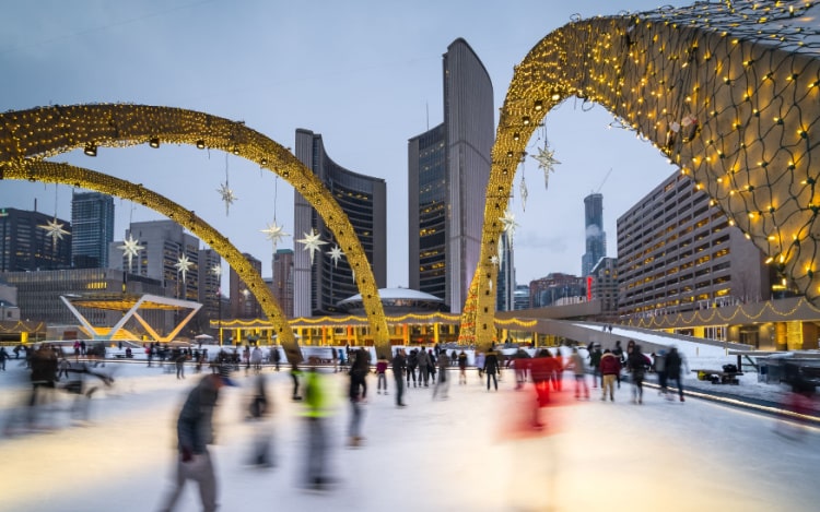 Several people ice skating on Nathan Phillips Square’s outdoor ice rink, with skyscrapers in the background.