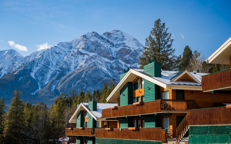 A partial view of a green and brown log cabin resort with balconies and snow-capped mountains in the background.