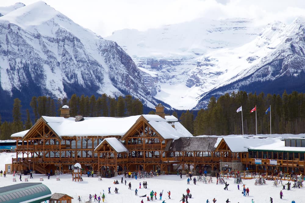 Wooden buildings with snow and flags on the roof. In front of the building are people wandering around; behind are tall trees and a view of the snowy mountains