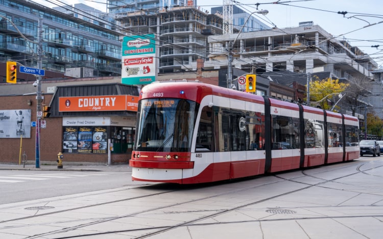 A red and white train car riding through a main street in Leslieville. There are multi-storey buildings in the background and a store with an orange sign.