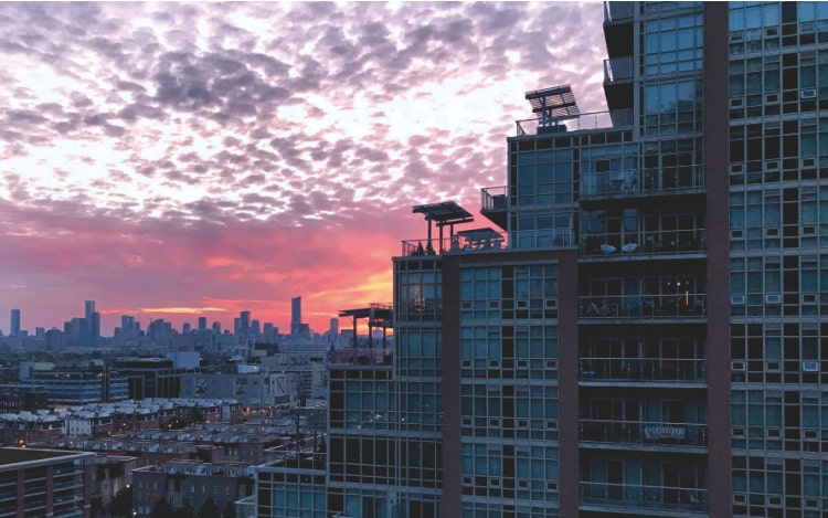 A partial view of a tall apartment building, with a view of the Toronto skyline at sunset. The sky is pink and orange.