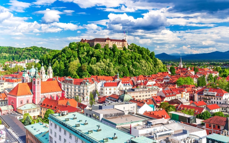 An aerial view of Ljubljana, with the tops of red, blue and white buildings visible. There is a green hill with a building perched at the top in the distance.