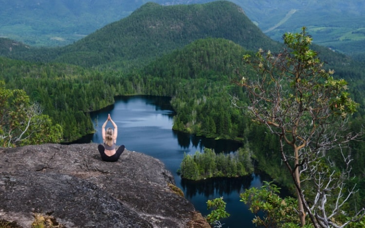 A woman meditates on a grey rock overlooking a forest and lake.