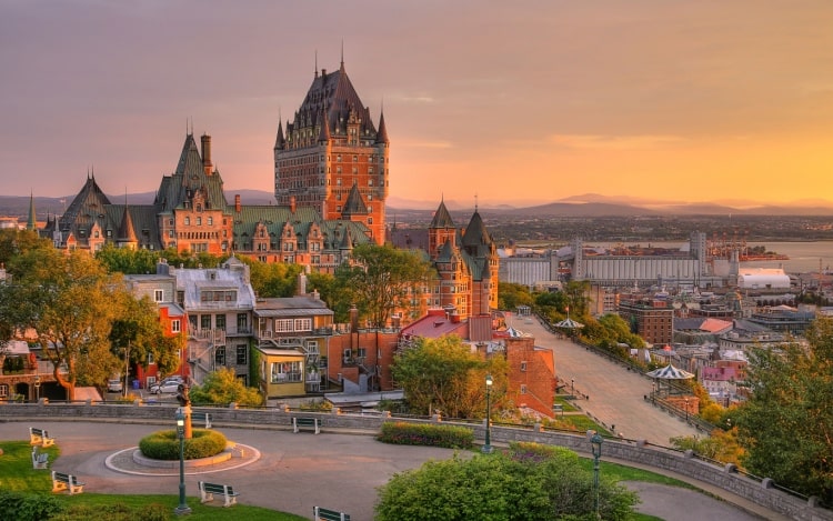 A town centre at dusk with a tall castle-like building towering above other buildings, a winding road, and a clear yellow-tinted sky.