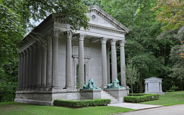 A large grey building with grey pillars and two green lion statues on either side of the stairs leading to the entrance. The building is under tree cover.