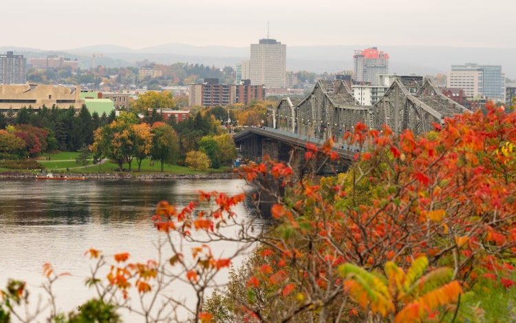 Ottawa in Fall with red, green and orange leaves in the foreground. There’s a grey river running under a bridge in the centre of the image.