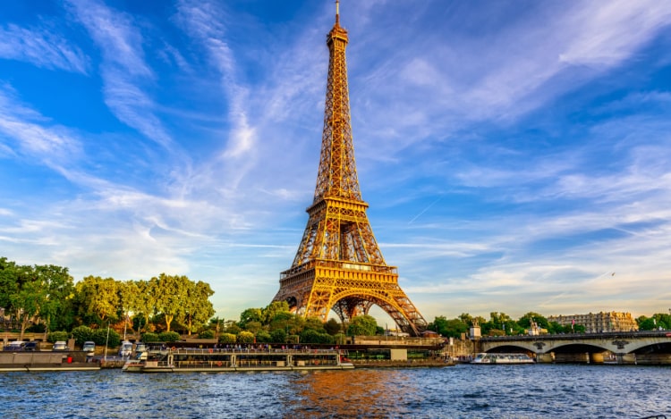 A view of the Eiffel Tower from the River Seine under a blue sky and wispy clouds.