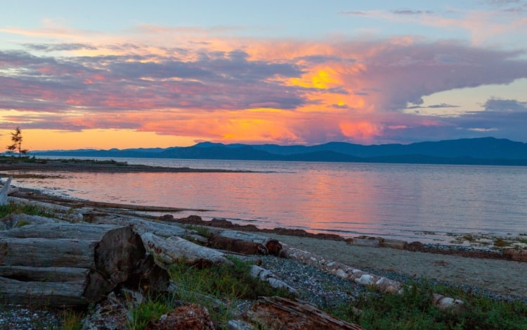 View of water from the coast in Parksville at sunset, with logs on a rocky beach, still water and an orange, pink and blue sky.