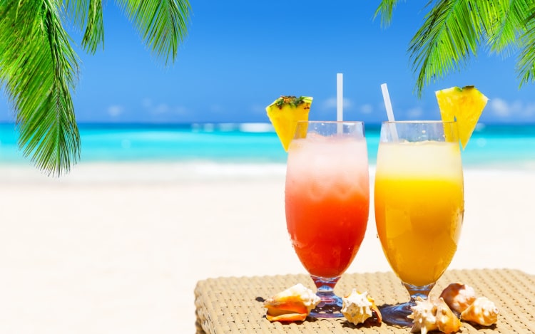 Two glasses; the left one is filled with red tropical juice, the right yellow. Both have ice cubes, a slice of pineapple stuck on the rims, and a straw.