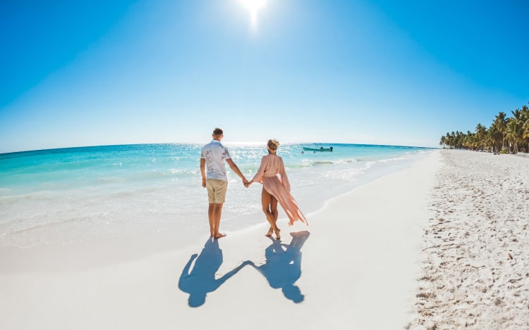 A white couple holding hands on a beach, facing away from the camera.