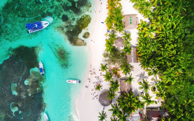 An aerial shot of a coastal part of Punta Cana. On the left is the clear turquoise water with 3 blue and white boats floating on it.