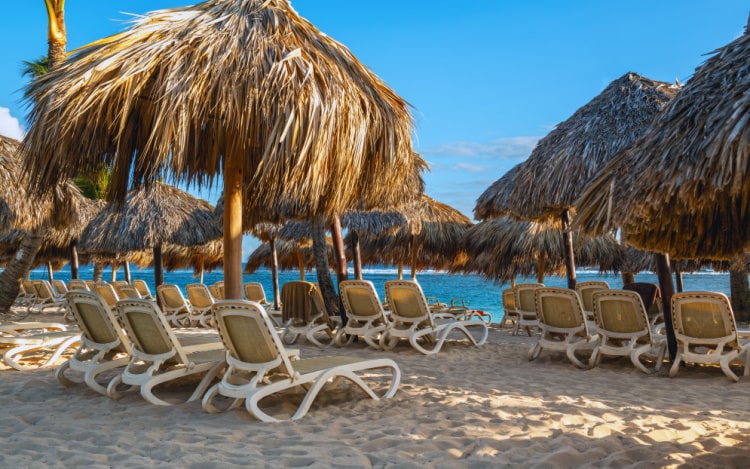 Several beige and white lounge chairs and straw umbrellas set up on the beach by the sea, which can be seen in the background.