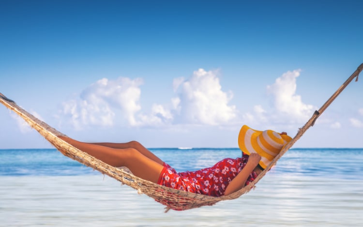 A woman wearing a red floral mini dress and a yellow and white striped sun hat is sleeping in a hammock with her face covered. The sea and sky are visible behind her