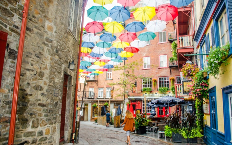 A side street in Quebec City with several coloured umbrellas suspended between the buildings and a woman walking away.