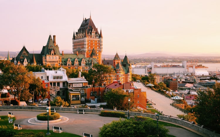 Quebec city in Fall under a clear pastel pink sky. In the foreground of the image is a road, in the centre there are several buildings.