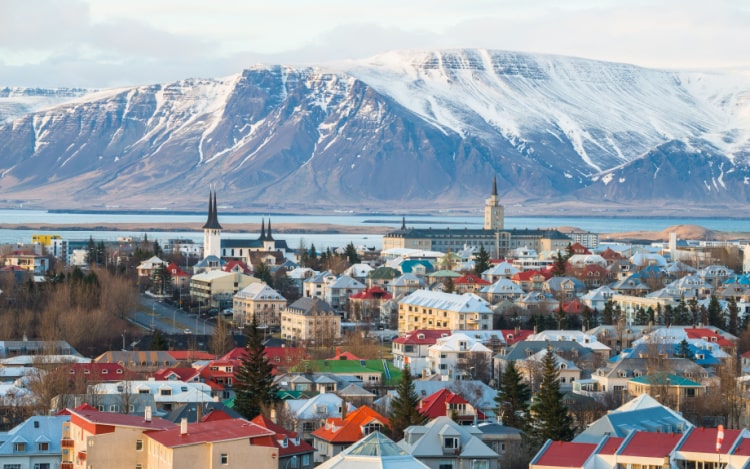 A view of Reykjavík in winter, with snow-topped buildings, blue water and a snowy mountain.