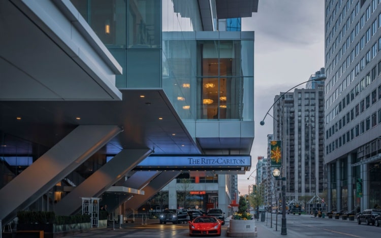 The entrance of Toronto’s Ritz-Carlton hotel underneath a glass building on a main road, with other white skyscrapers visible.