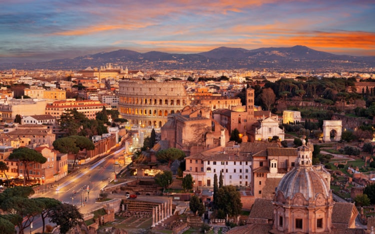 Rome at sunset, with a view of the main road, the Colosseum, and mountains in the distance under an orange and blue sky.