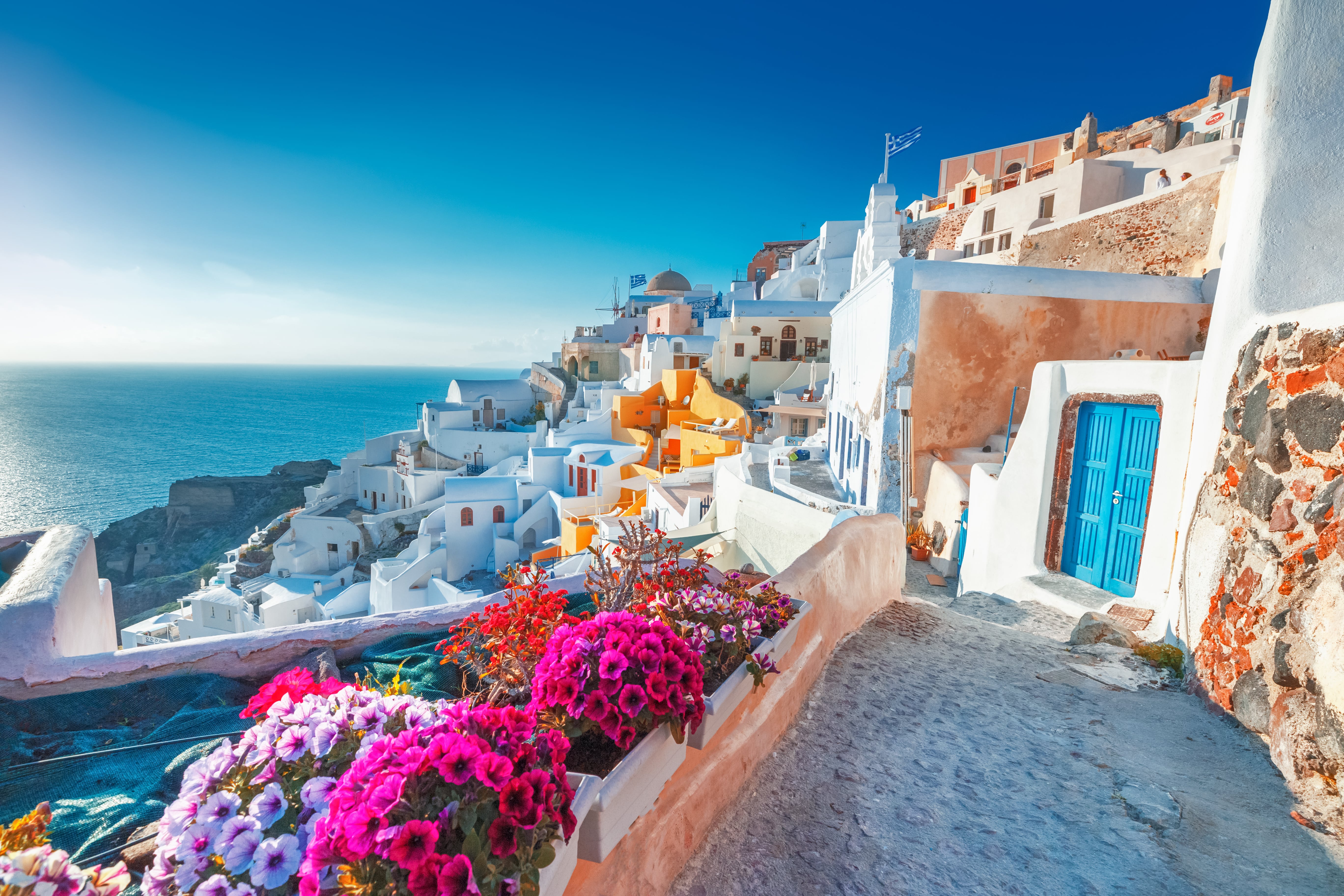 Picture of classic white Greek architecture adorned with pink and purple flowers overlooking the coast
