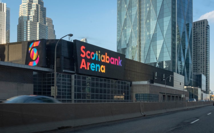A partial view of the Scotiabank Arena taken from a road, with a multicoloured sign and tall buildings in the background.