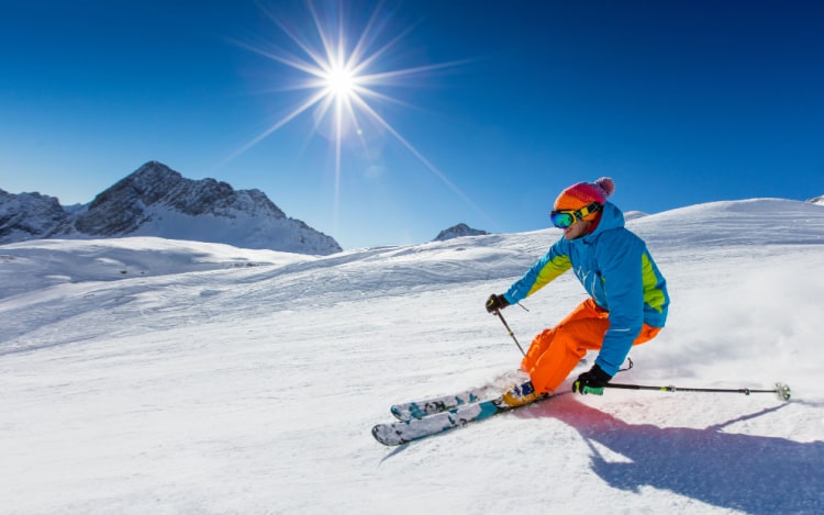 A man in blue and orange skiing gear and goggles skiing down a snowy mountain. The sky is clear, and the sun is shining brightly.
