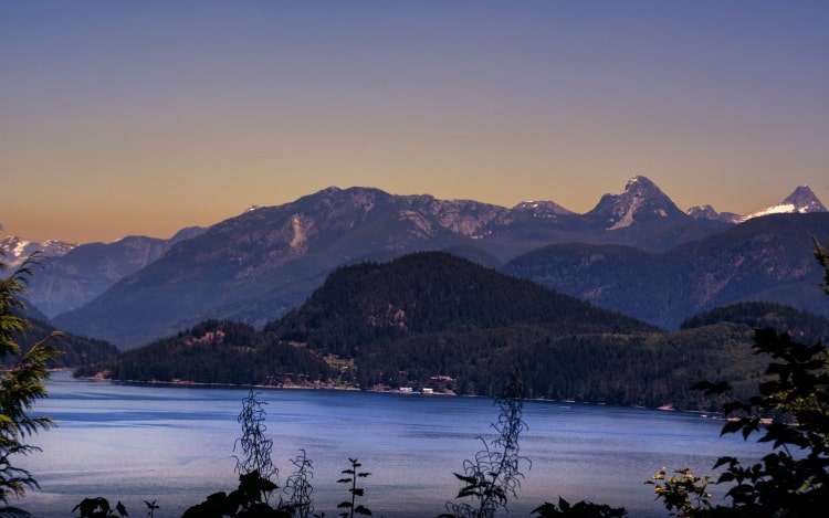 A still lake at dusk with trees and mountains visible in the distance.