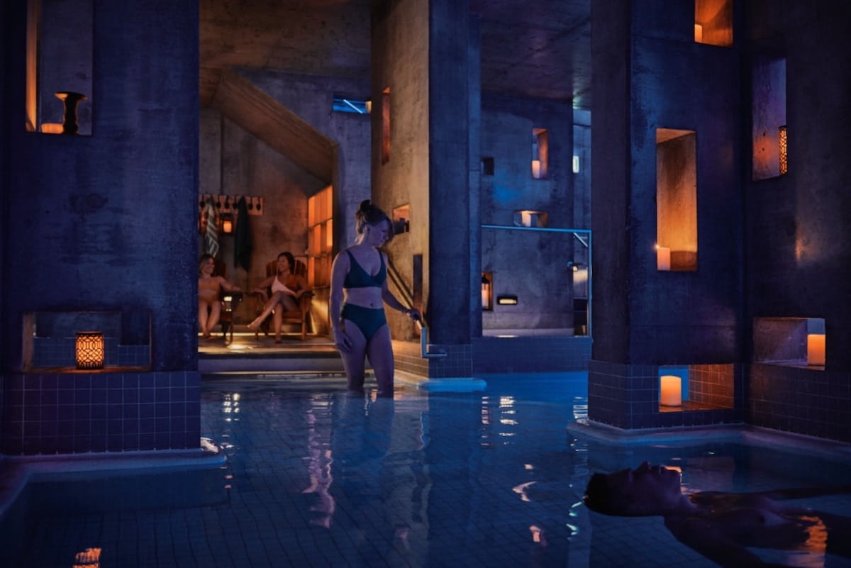 Dark blue and purple spa room with a person walking into the pool and two people sat on chairs in the background