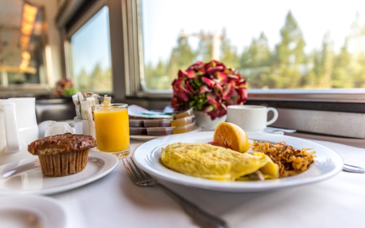 Breakfast in a first class train car, including an omelette, muffin, orange juice and fruit, with flowers and the train windows in the background.