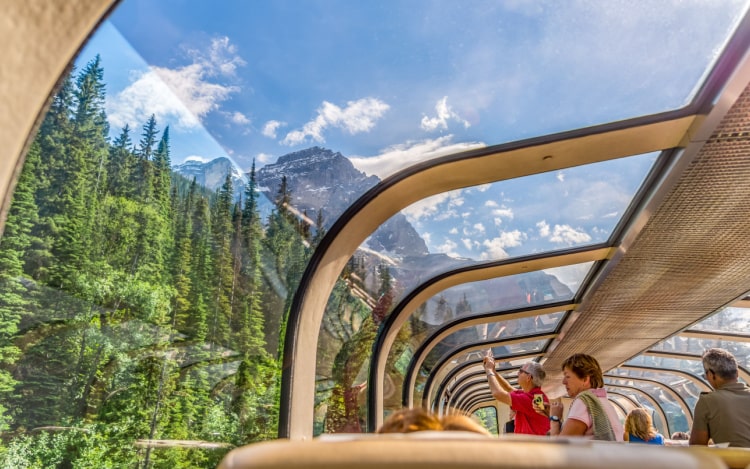 The inside of a train car with curved windows that extend to the roof, with trees outside, a blue sky and a mountain in the distance.