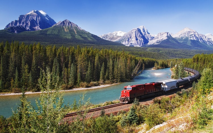 A red train runs past a blue river, green trees and snow-capped mountains.