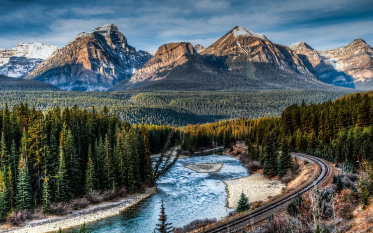 A train line winding past a river, green trees and mountains in the distance.