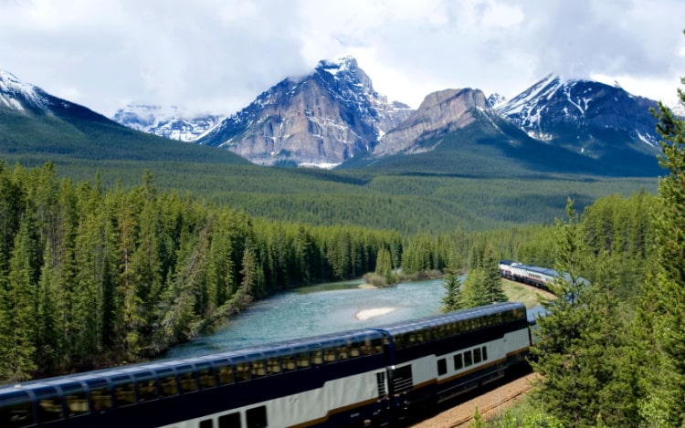 A parietal view of a blue and white train on tracks passing through trees by a river with mountains in the background.