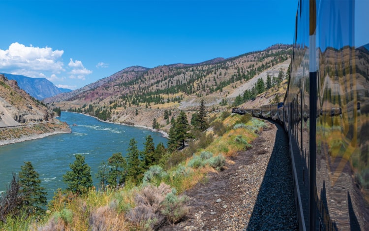 A view out the window of a moving train next to a blue river on a clear day.