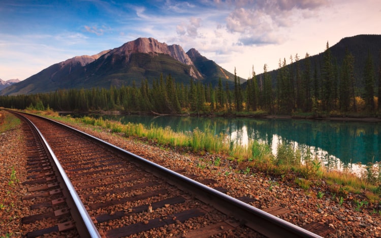 Train tracks over red dirt with a still river, trees, and a mountain to the right.