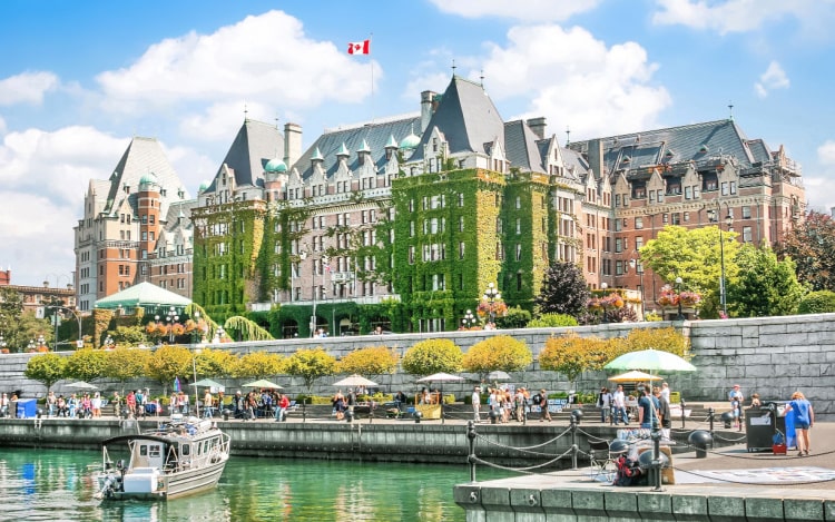 A view of the Inner Harbour of Victoria, Vancouver Island, with one boat in the still waters. It’s a clear day, and there is green ivy climbing the large building in the background.