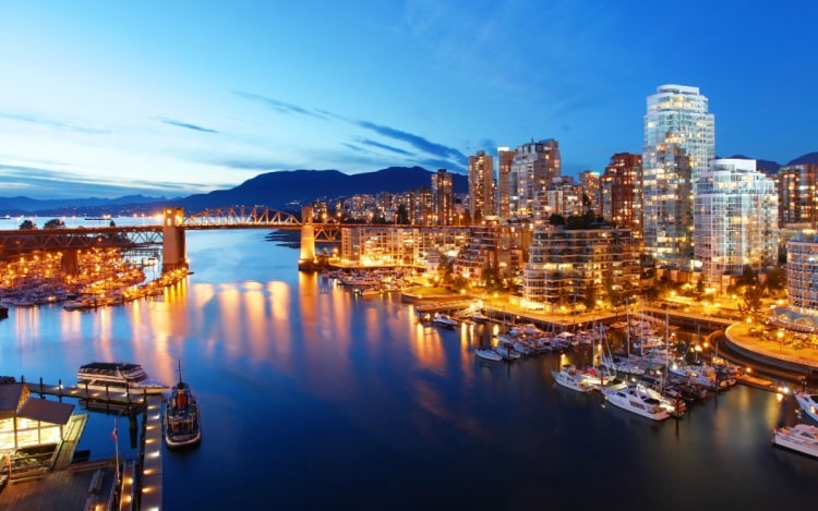 The city of Vancouver from the river at dusk, with all the buildings illuminated, boats in the water, and mountains in the distance.