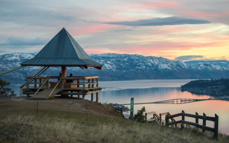 Sunset over Lake Okanagan with a hut overlooking the water, pinks and yellows being reflected in the still water and snowy mountains in the background.