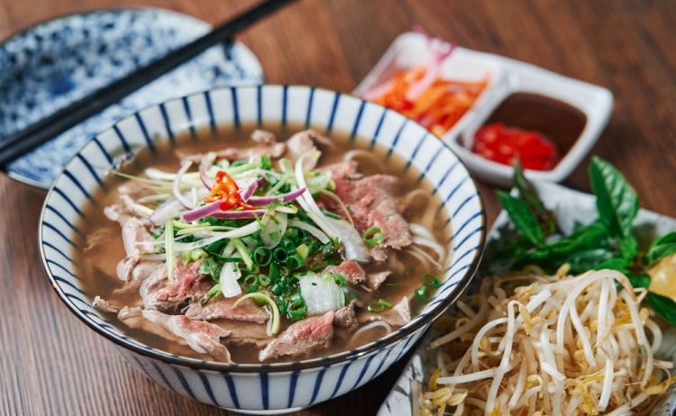 A black and white striped bowl filled with clear soup, noodles, strips of beef and topped with vegetables. To the right of the bowl is a serving of beansprouts, along with chopsticks and more green and red toppings out of focus.