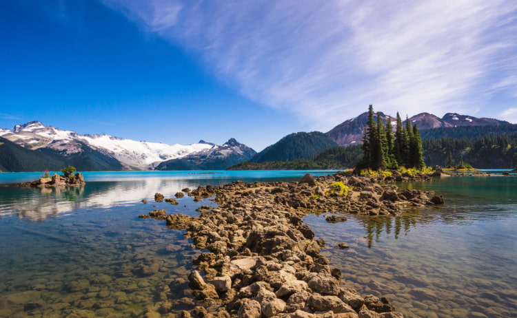 Crystal-clear waters of a lake with a rocky path in the centre leading to a small island of trees, with snowy mountains in the background