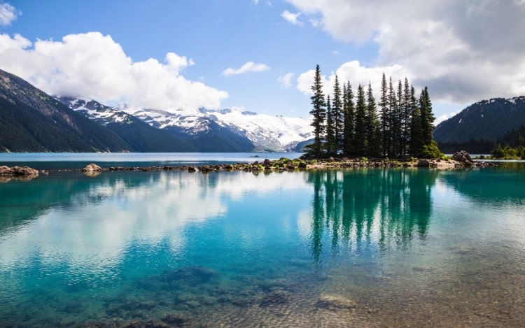 A still blue lake with a small island of trees off to the right and snow-capped mountains in the background.