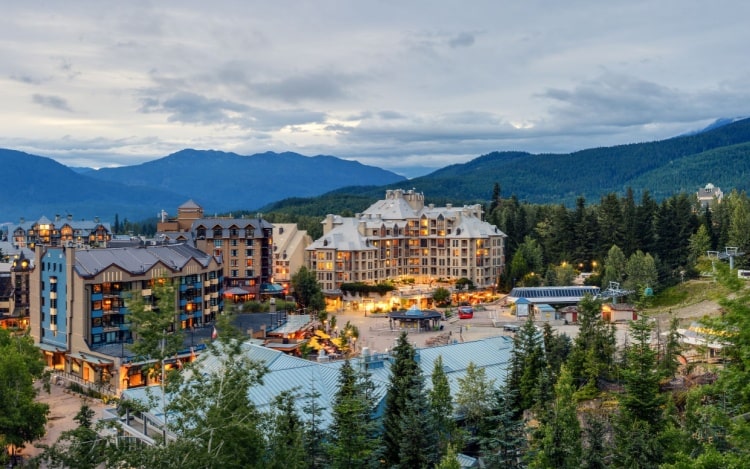 Whistler village on an overcast day, with lights on in the three visible resort buildings, green trees surrounding the village and mountains in the background.