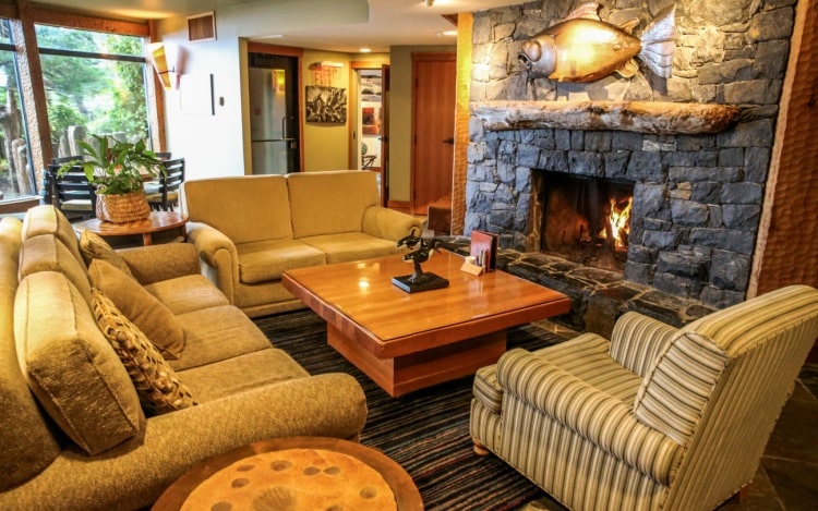 A resort’s lounge, with multiple beige sofas surrounding a square brown coffee table and a grey stone feature wall with a lit fireplace inside.